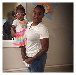 Black woman holds young black child who wears pink framed glasses and a butterfly dress.