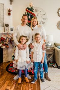 smiling father, mother, sister, brother standing in front of Christmas tree.