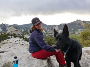 Image of Michelle (left) and her dog (right) sitting on a rock during a cloudy day. Mountains and trees in the background.