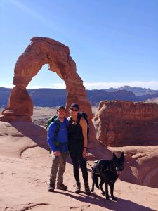 Jason (left) and Michelle (right) outside smiling at the camera. To Michelle's side, is her guide dog, Ryder. The background is of a blue cloudless sky and rust/orange rocks. Left middle of image is a rock arch.